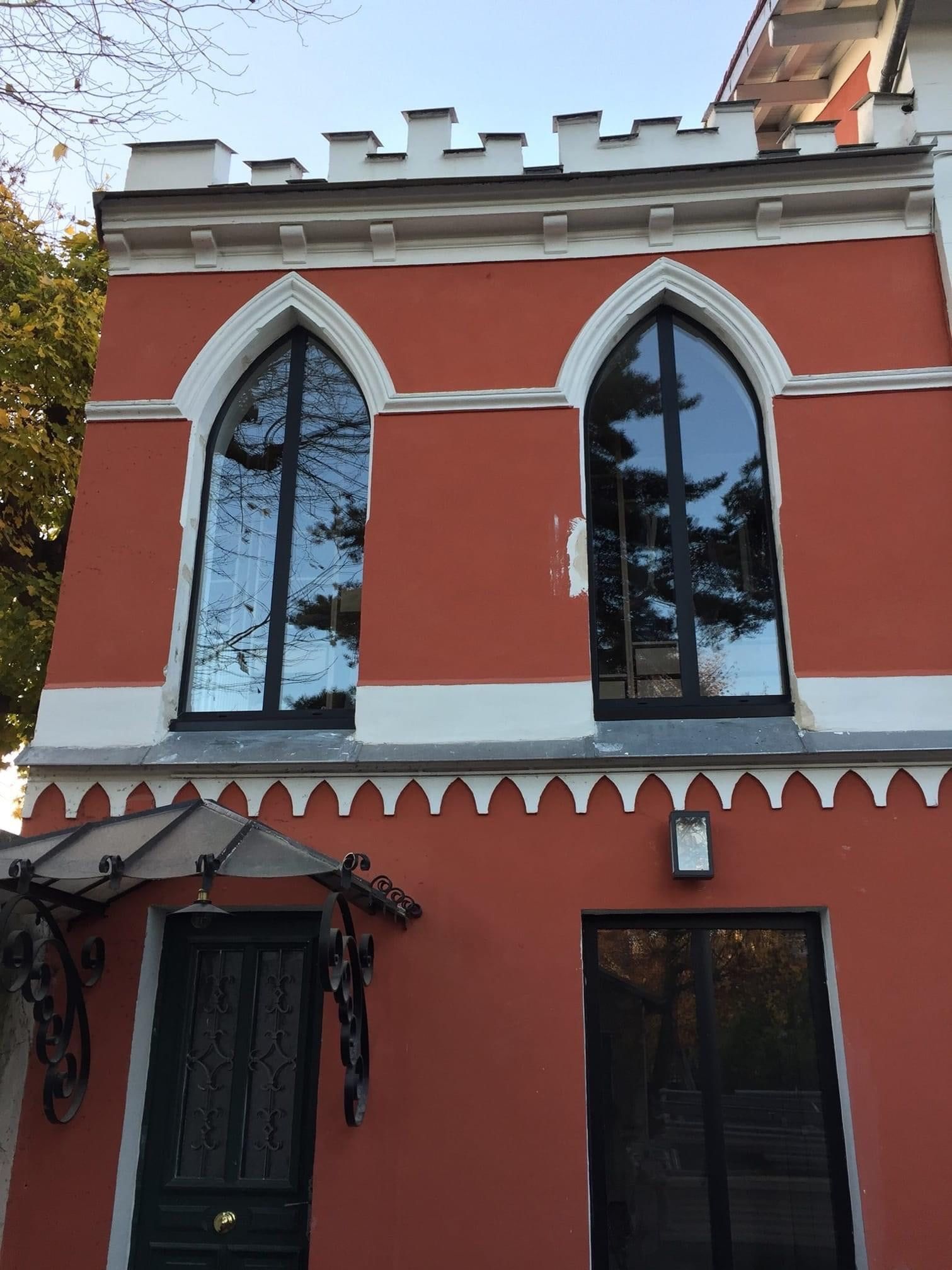 Façade rouge d'une maison de style néogothique, deux fenêtres aluminium en arc brisé sous un couronnement crénelé