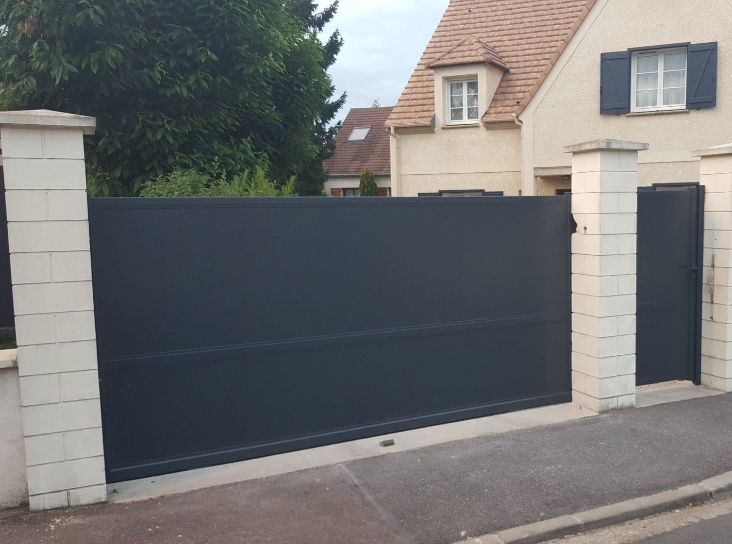 Portail et portillon aluminium gris anthracite à remplissage plein, entre des piliers maçonnés clairs, devant une maison