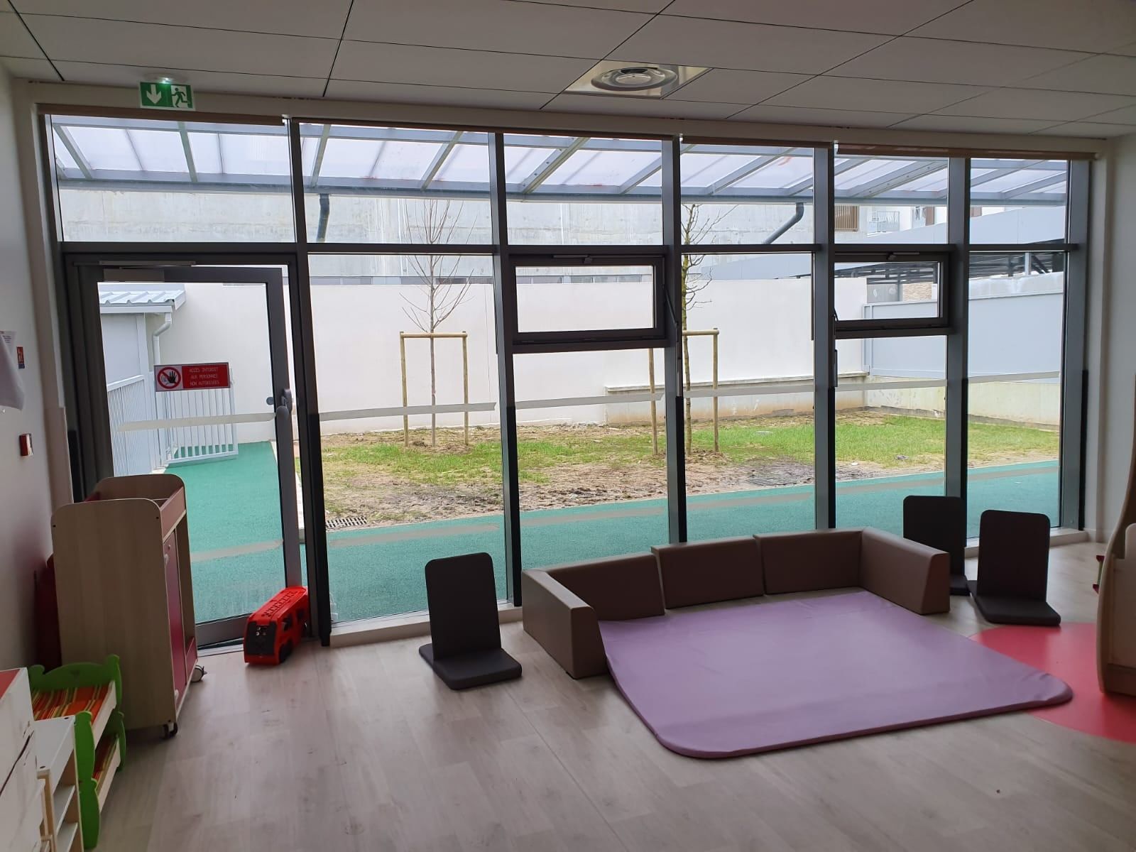 Salle d'activités de crèche vue de l'intérieur ; à gauche, la porte vitrée aluminium ouverte sur la cour de jeux